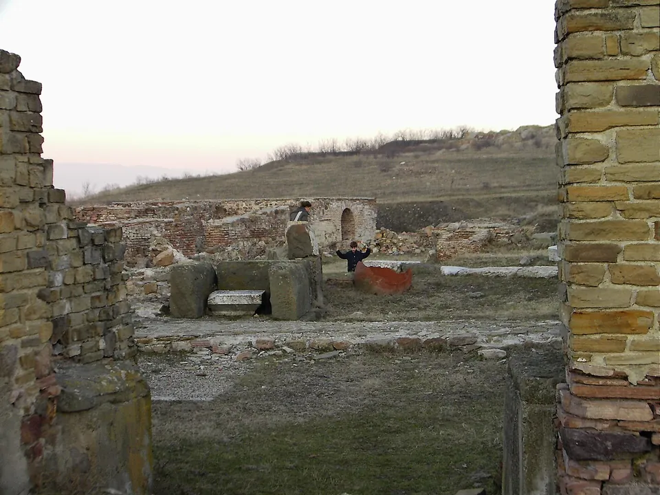 Excavated Roman walls of pale squared stone with brick courses framing a view of foundations, a round-arched doorway and a grassy hill beyond, with a child standing among the ruins.