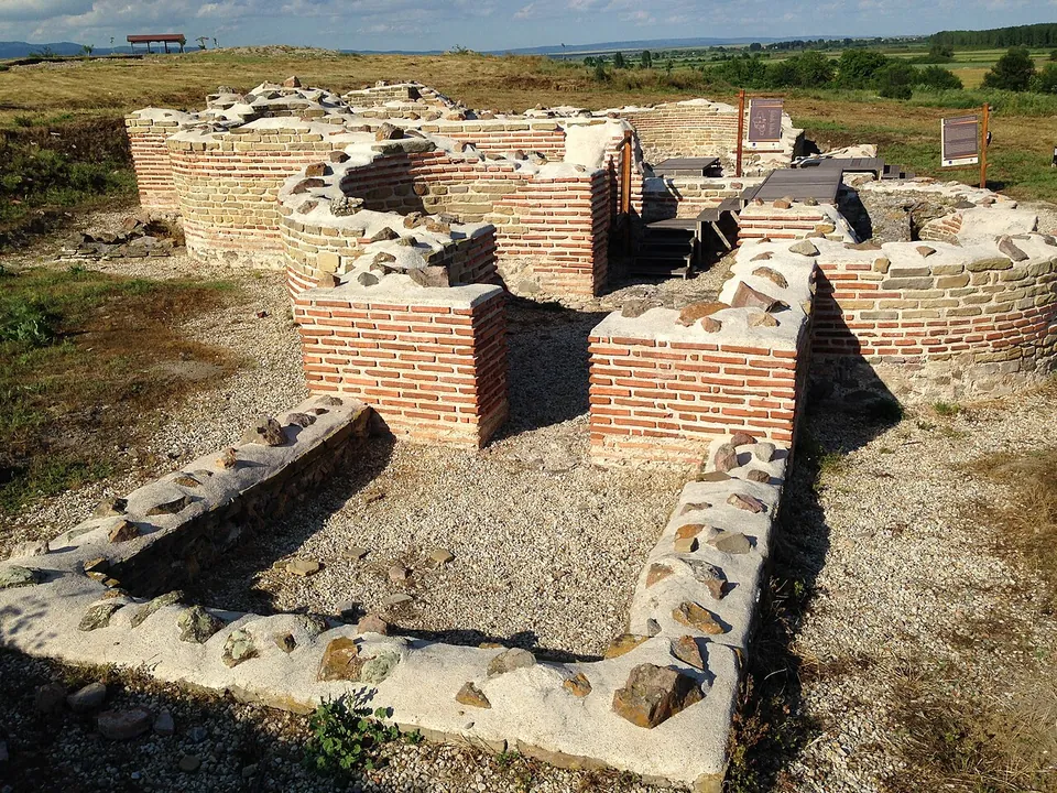 Restored low walls of stone and red brick forming rounded and square rooms on gravel, with information boards and open grassland beyond.