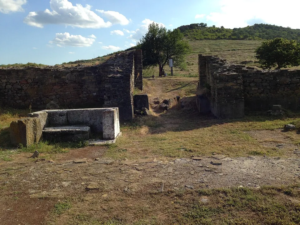 The opening of a gate between two thick stretches of stone wall, a large carved stone block in front of it and a grassy, wooded hill behind.