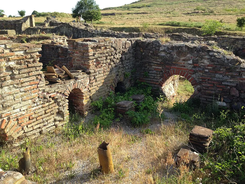 Ruined walls of brick and stone with several round brick arches at floor level and the stumps of clay pipes in the ground in front.