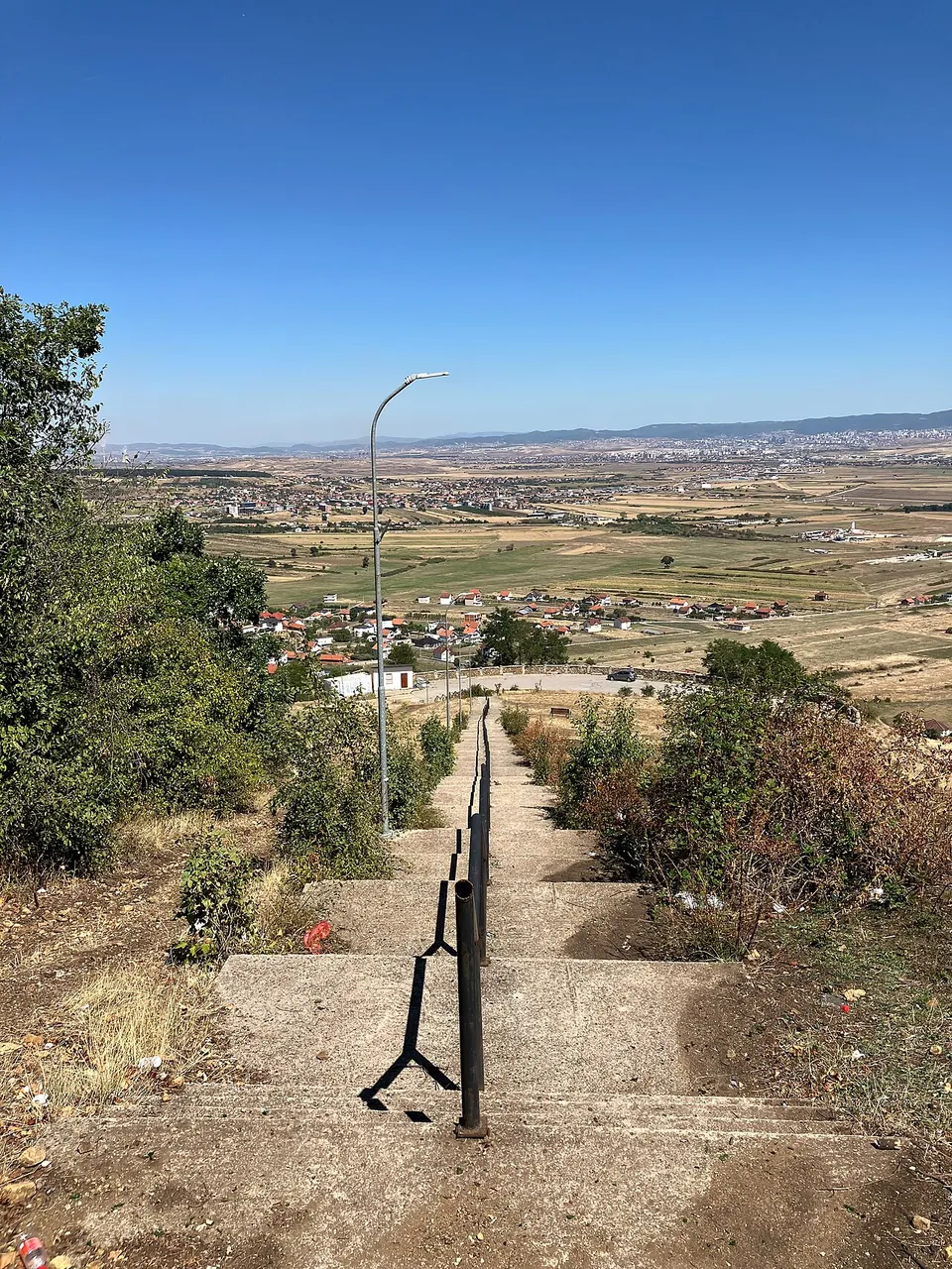 A long flight of concrete steps with a metal handrail and a street lamp running down a scrubby hillside, with villages and farmland spread across a wide plain below.