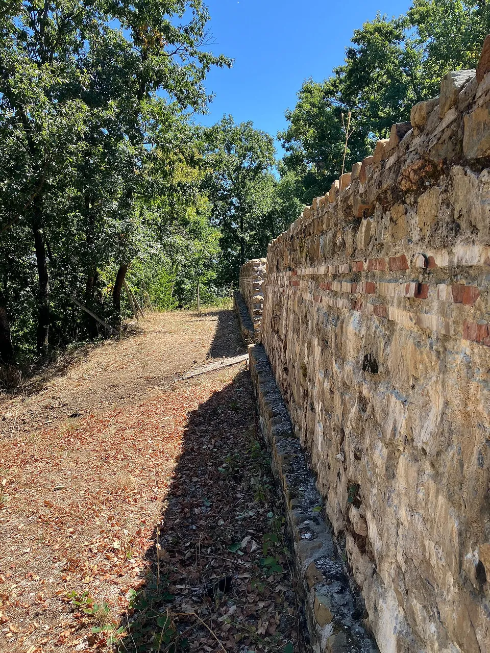 A tall wall of roughly shaped stone laid with thin horizontal bands of red brick, with a leaf-covered path running along its foot among trees.