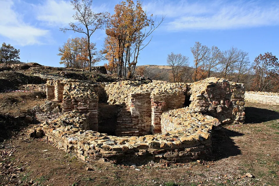 An excavated structure of rubble stone with piers built of flat red bricks, curving round an open interior, on bare ground with autumn trees behind.