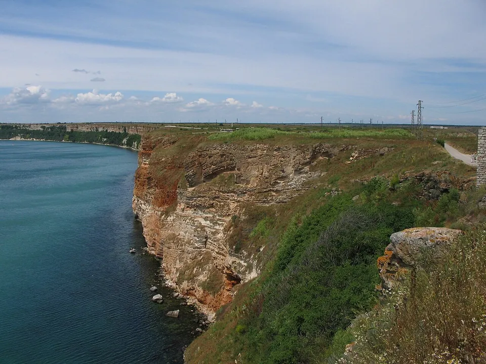 Sheer layered cliffs of ochre and red rock falling to clear turquoise water along a long straight coast, with green grass on top.