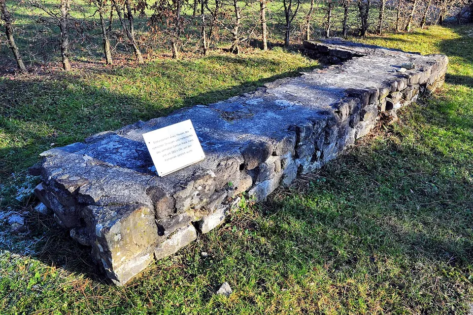 A short, thick length of rough stone wall lying in a grassy slope, with a small white plaque fixed on top and bare shrubs behind.