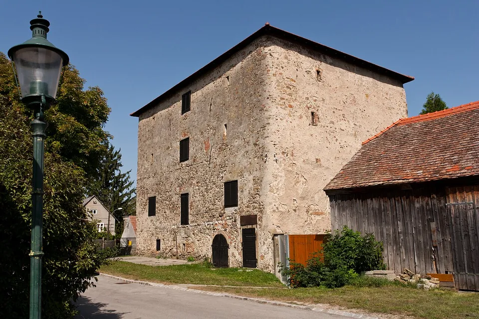 A tall, plain three-storey building of rough stone with a steep dark roof and small shuttered windows, beside a timber barn with a tiled roof, an old street lamp in front.