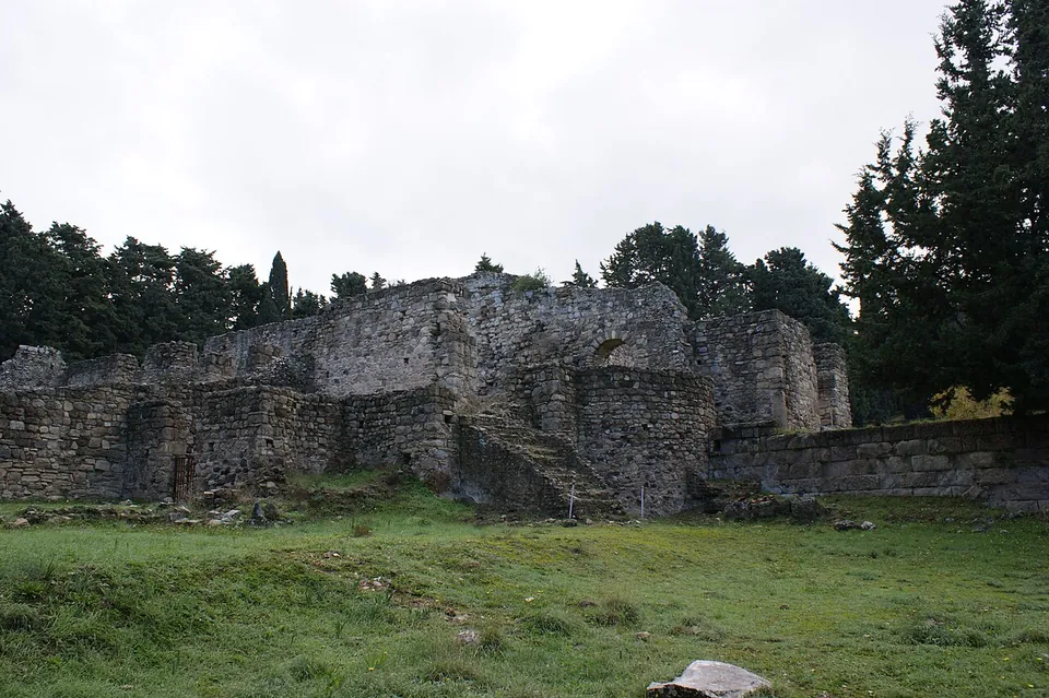 The ruined walls of a building of rough stone and mortar, with arched openings and a stair, standing on a grassy slope against dark pines and a grey sky.