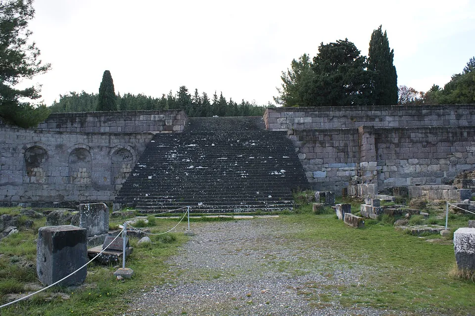 A broad flight of stone steps rising up the middle of a long retaining wall of squared blocks, with round-headed recesses set into the wall to the left, a gravel path and scattered stone bases in the foreground, and pines above.