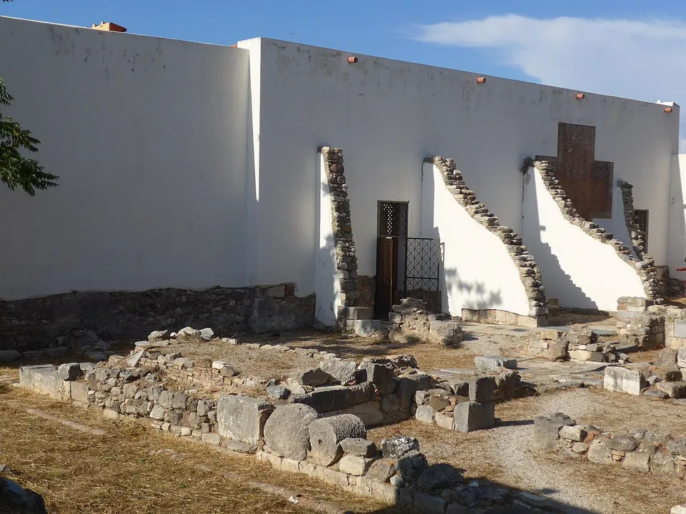The plain whitewashed outer walls of the rebuilt house, with original stone wall ends and a doorway, above excavated foundations in the foreground.