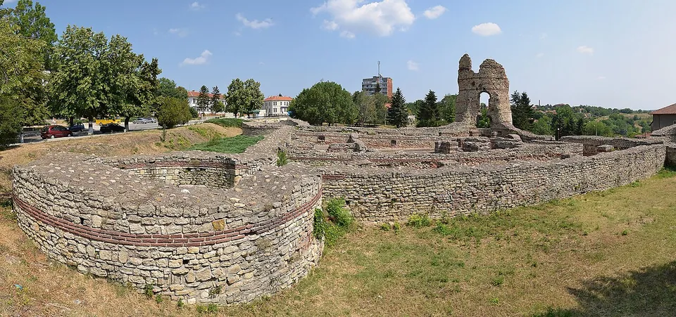 A panorama of a square Roman fort of stone with bands of brick, a round corner tower in the foreground and a tall ruined tower with an arched opening rising at the far corner, on a grassy slope among trees and town buildings.