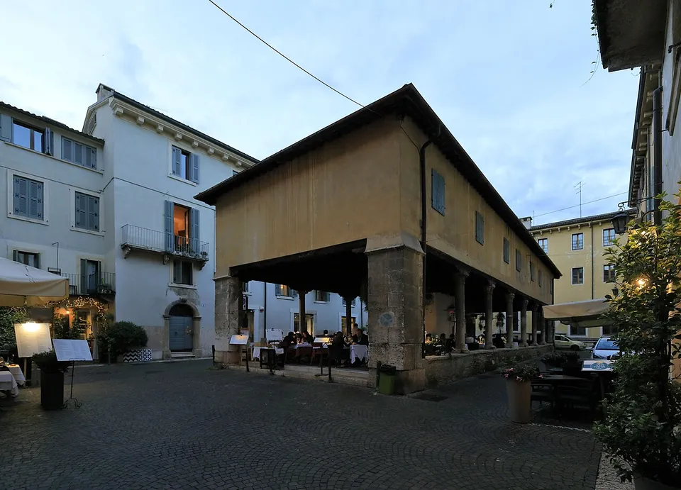 A long two-storey building of ochre plaster raised on a row of stone columns over an open ground floor, with restaurant tables beneath it, standing in a small paved square at dusk.