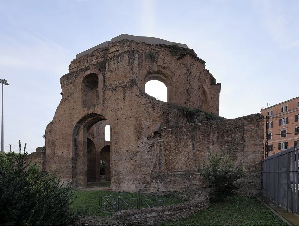 A tall ruined brick building with a broken upper drum pierced by arched windows, a large arched opening at ground level and lower walls around it, on a lawn with a modern apartment block to the right.