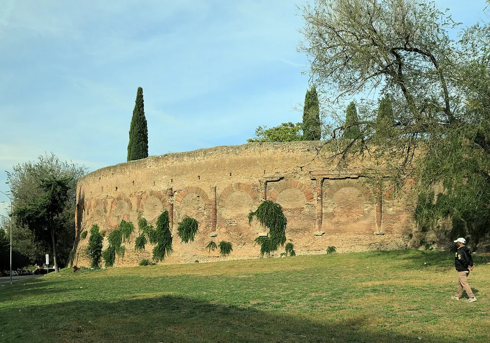 The curve of a brick wall across a wide lawn with cypresses rising behind it, ivy hanging from the blocked arches of its lower storey and a man walking on the grass in front.
