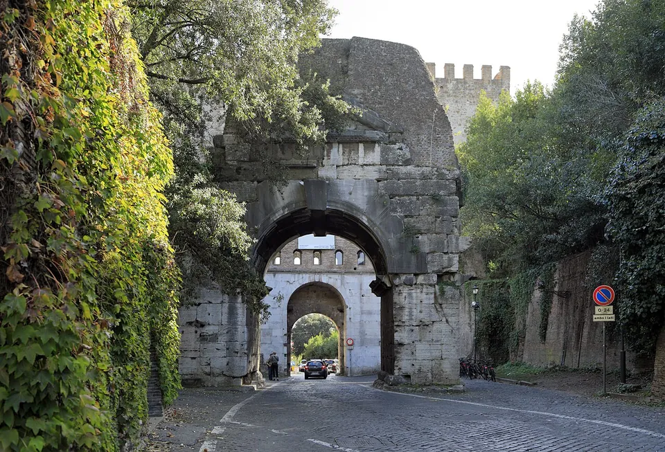 A weathered single stone arch spanning a cobbled road between ivy-covered walls, with the pale archway of a gate visible through it and the top of a crenellated tower behind.