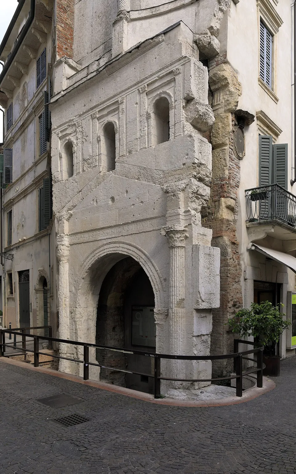 The surviving half of a pale stone gate front built into the corner of a later plastered house. A round arch at street level is framed by fluted half-columns with leafy capitals under a line of carved lettering and a low pediment; above are small arched windows between pilasters and, higher still, a broken upper storey. Along its right-hand edge the rough stone gives way to older red brickwork. A low railing runs round the foot of it on a cobbled street.