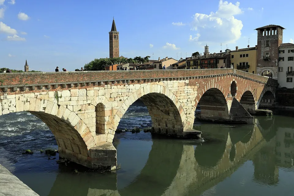 A bridge of round arches over a broad green river, the two nearest arches built of large pale stone blocks and those beyond of brick, with a brick bell tower and houses on the far bank.
