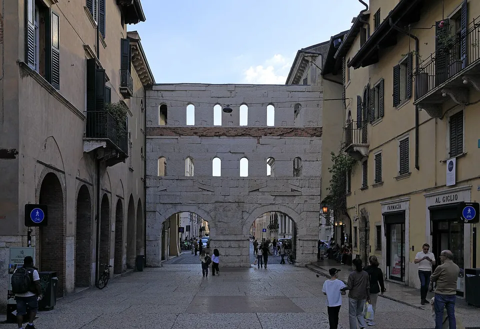 A paved shopping street lined with shuttered houses, closed at its far end by a flat stone wall with two arches at the bottom and two rows of plain arched openings above, through which the sky shows.