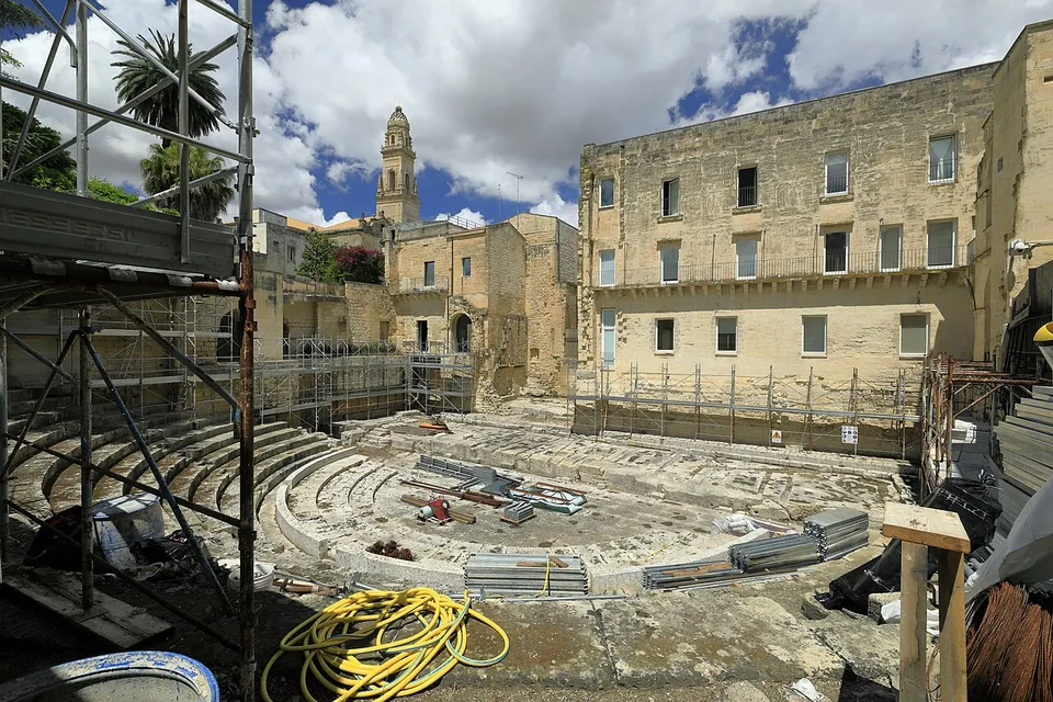 The theatre seen past scaffolding, with metal frames, hoses and building materials lying across the seating and orchestra, and a three-storey stone building and a bell tower behind.