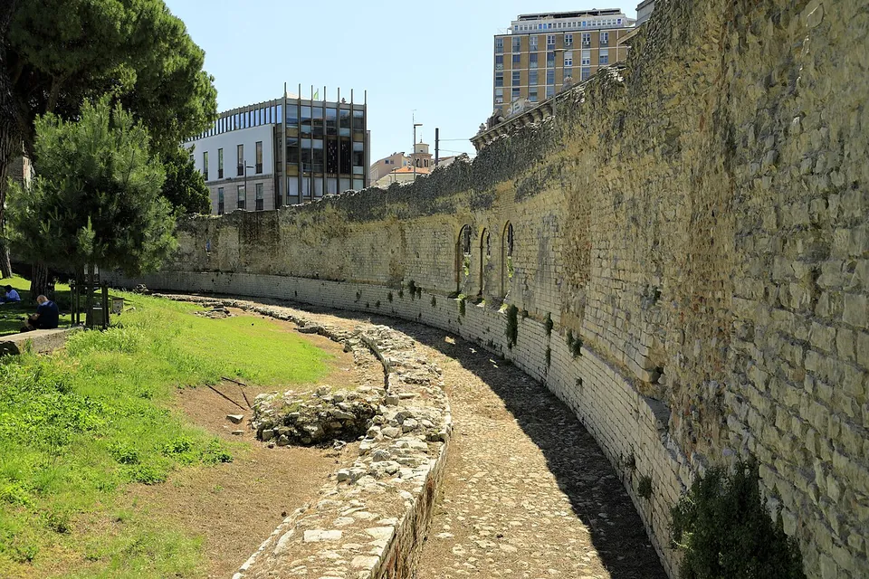 A long curving wall of pale limestone blocks, its upper part ragged and overgrown, with three small arched openings near its far end, running round a sunken grassy strip with low foundations, modern buildings above it.