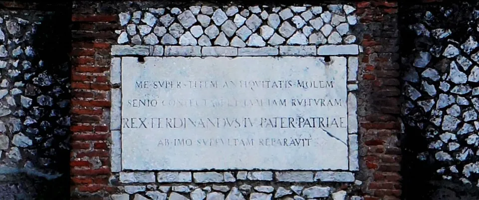A white marble plaque with a carved Latin inscription in capitals, among them the words REX FERDINANDVS IV PATER PATRIAE, set into a wall of small diamond-shaped stones framed by red brick.