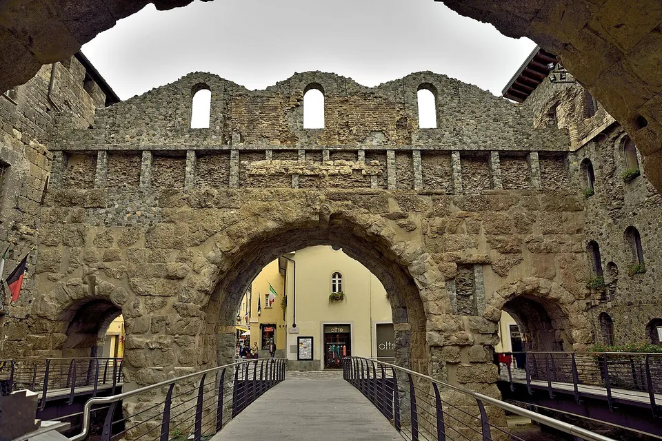Seen from under an arch, a footbridge with metal railings leads across a sunken area towards a large round stone arch flanked by two smaller ones; above them runs a band of stonework and a ruined upper wall with three arched window openings, with a town street visible through the central arch.