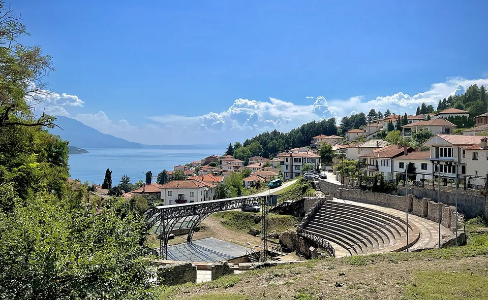 The stone seating of the theatre among the red-roofed houses of the old town, with the lake and mountains beyond under a summer sky.