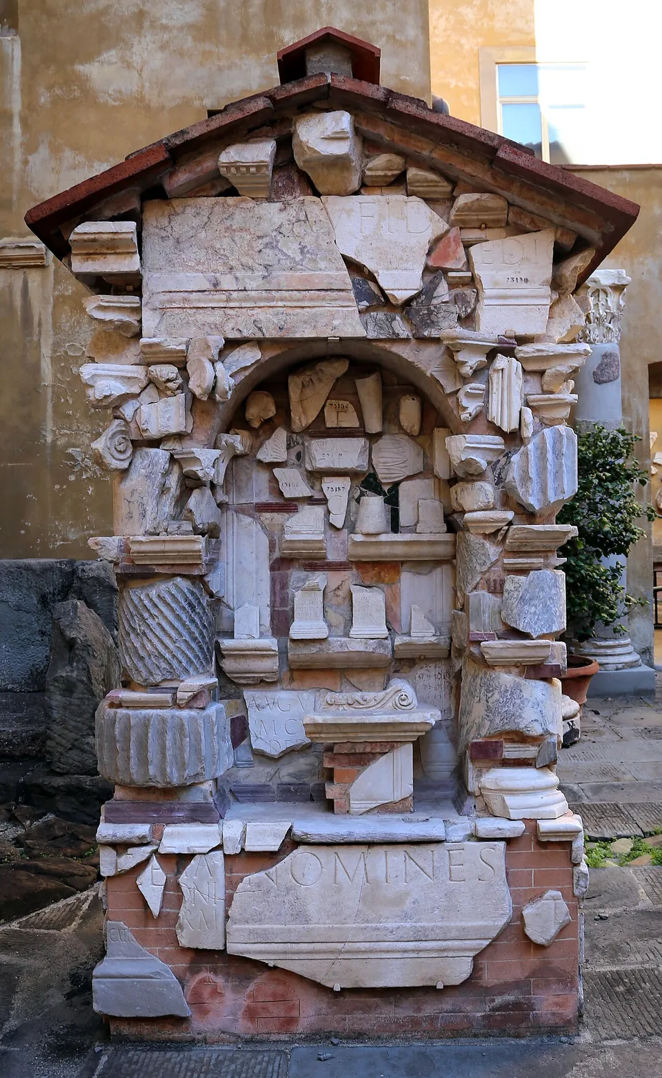 A small gabled structure with a tiled roof, built up from dozens of white marble fragments - mouldings, fluted column pieces, carved slabs - with a slab at the bottom carrying a few large Latin letters.