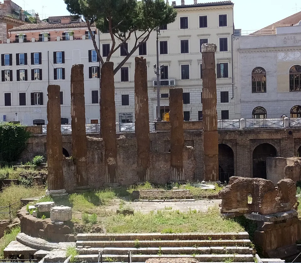 Six tall, weathered column shafts standing in a curve on a round stone podium, with a flight of steps in front and a brick arcade and street railing behind.
