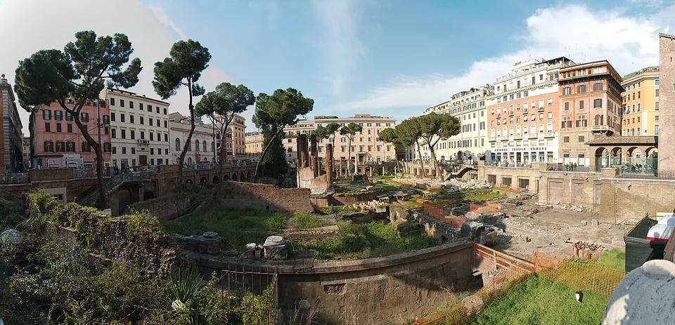 A wide panorama of a sunken rectangle of ruins in the middle of a city square, with grass, low walls and column stumps below street level, umbrella pines along the edge and pastel-coloured apartment buildings all round.
