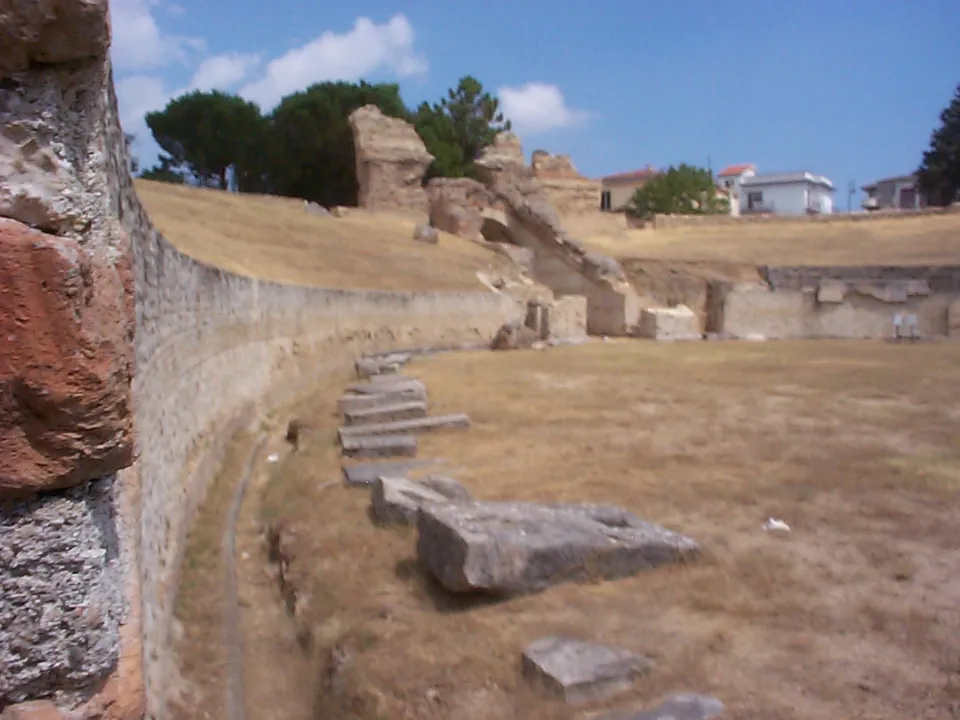 The curving stone wall round the arena, with large fallen blocks lying on the dry grass of the arena floor and ruined masonry on the slope above.
