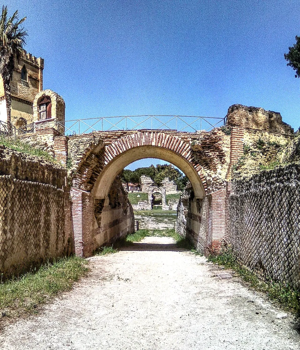 A gravel path running through a brick-arched passage between walls faced with small stones set in a diamond net pattern; a footbridge with a railing crosses above the arch, and ruins and trees show through it.