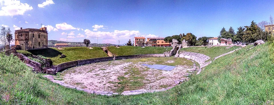 A wide panorama from the upper slope of the amphitheatre - an oval arena of pale bare ground ringed by a low wall, grassed banks rising round it, a few stretches of ruined masonry on the far side, and a brick building and modern houses beyond.