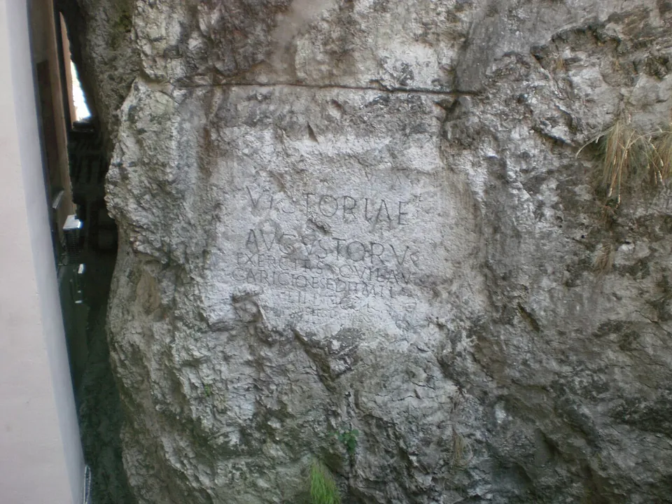 A face of pale, rough limestone rock with an inscription carved into a smoothed panel, wedged into a narrow gap beside the glass and white wall of a building on the left.