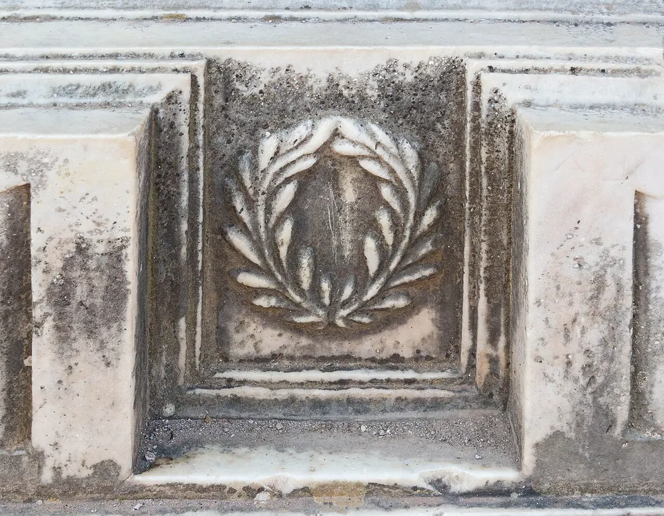 A close-up of weathered marble moulding with a laurel wreath carved in a square recessed panel.