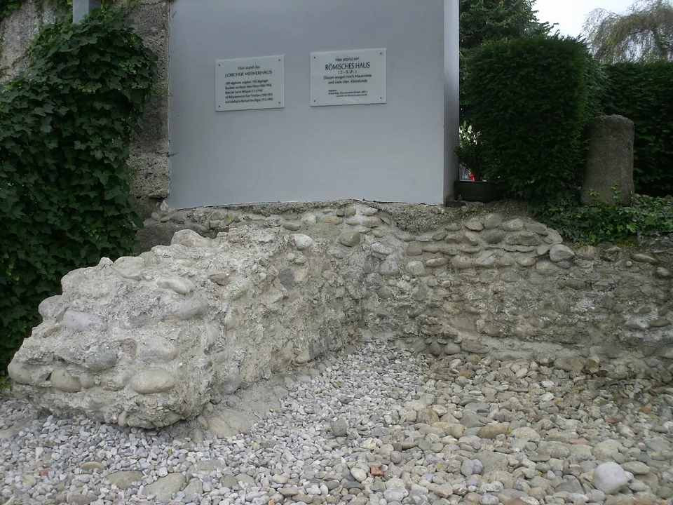 A low length of wall built of rounded stones set in mortar, laid out on gravel in front of a white panel with two small signs, between hedges.