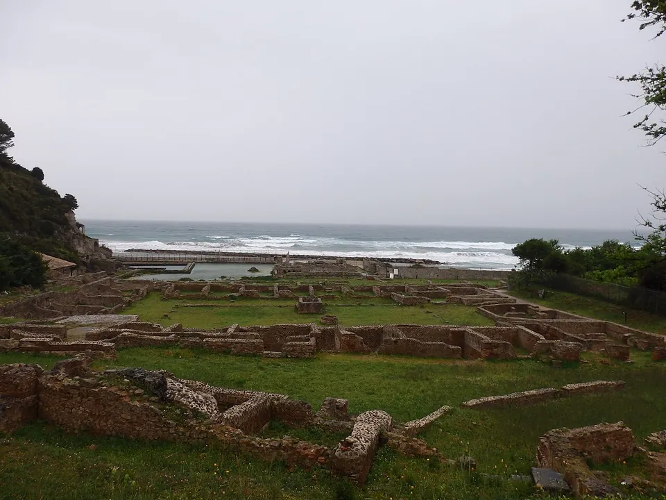 The foundations of many small rooms, walls of rough stone a few courses high, spread across a lawn towards the sea, with a wooded headland on the left.