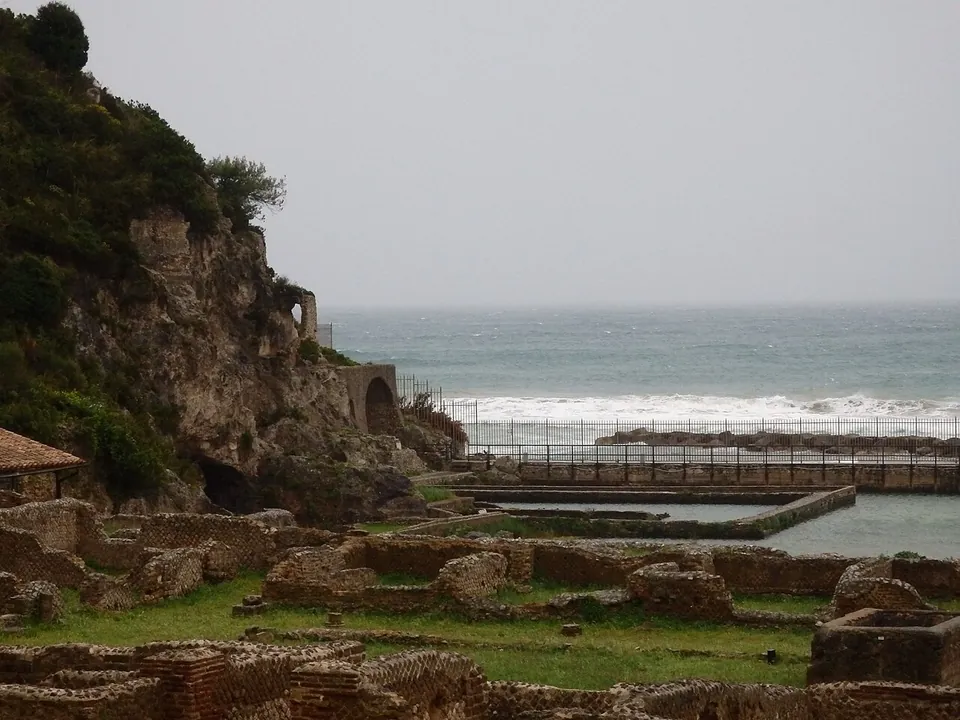 Low ruined walls of stone and brick in the grass in the foreground, beyond them a long stone-edged pool and the sea breaking on the shore, and on the left a rocky cliff with an arched opening at its foot.