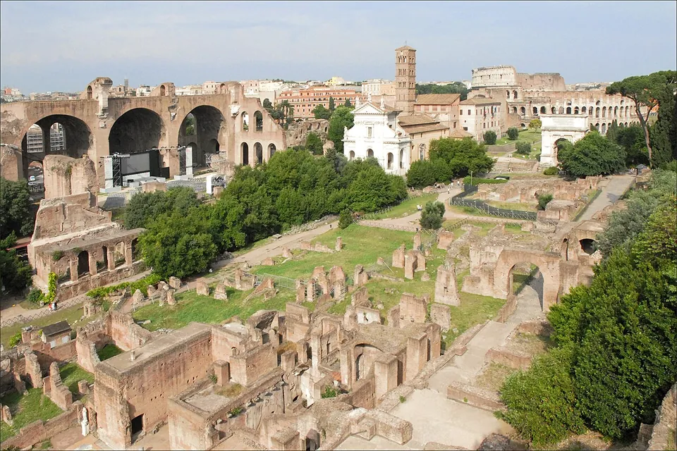 A view down over the eastern half of the Forum from a height, low brick and stone ruins among trees and lawns, with three huge brick vaults on the left, a white church front and a bell tower in the middle, and the curved wall of the Colosseum on the horizon.