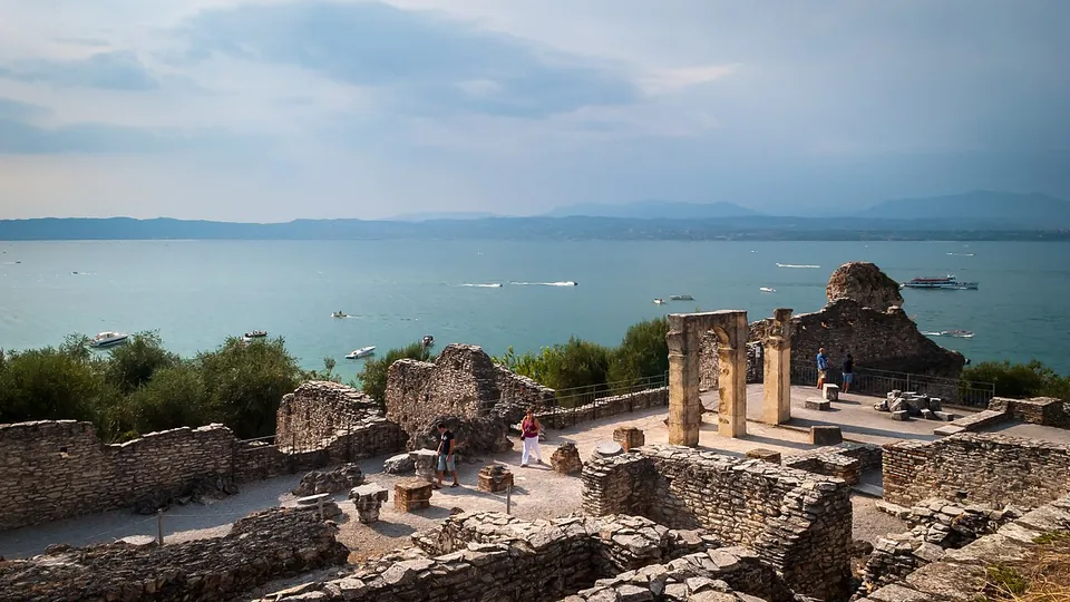 Low stone walls and the bases of rooms step down a slope in the foreground; beyond them two stone pillars carrying a lintel and a rough block of masonry stand at the edge of the site, with the lake, motorboats and the far shore behind.