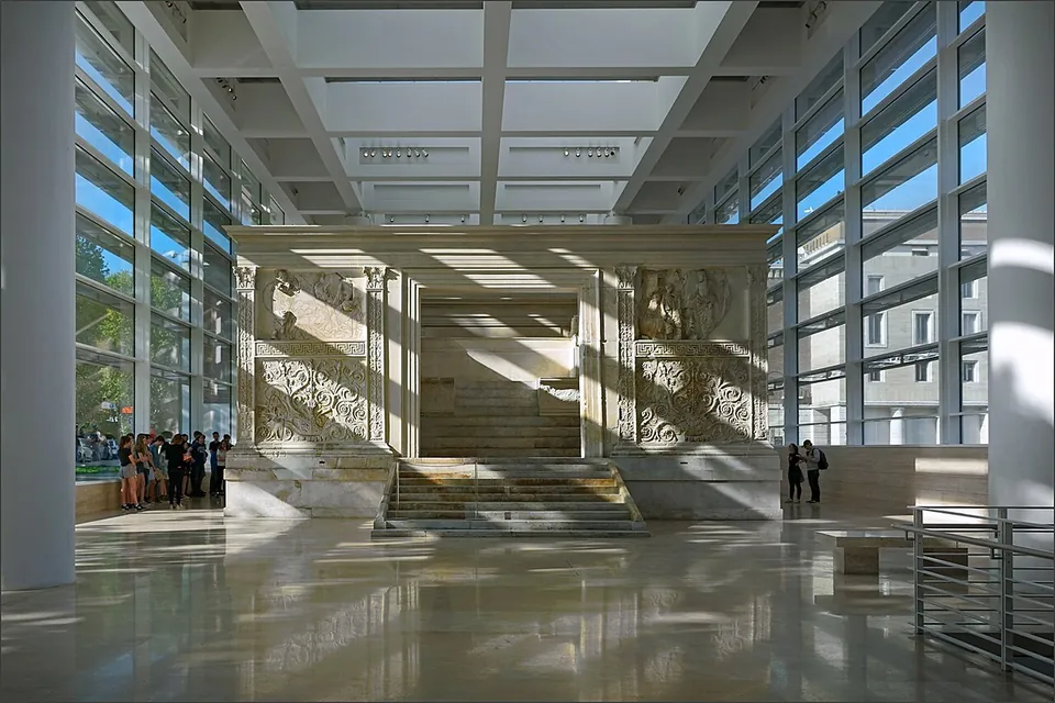 A long, bright hall with floor-to-ceiling glass walls and a grid of skylights; the pale marble enclosure of the altar stands in the middle on its steps, with small groups of visitors on either side.