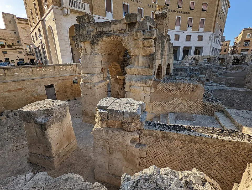A stone arch and square piers of large pale blocks, with lower walls between them faced in a net-like pattern of small square stones set on the diagonal, and modern buildings behind.