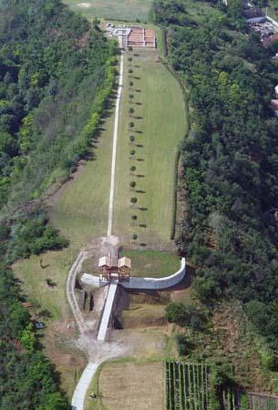 An aerial view along a long, narrow grassy plateau between wooded slopes, with a path running its length, a timber gate structure and a curved stone wall in the foreground and a walled building outline at the far end.