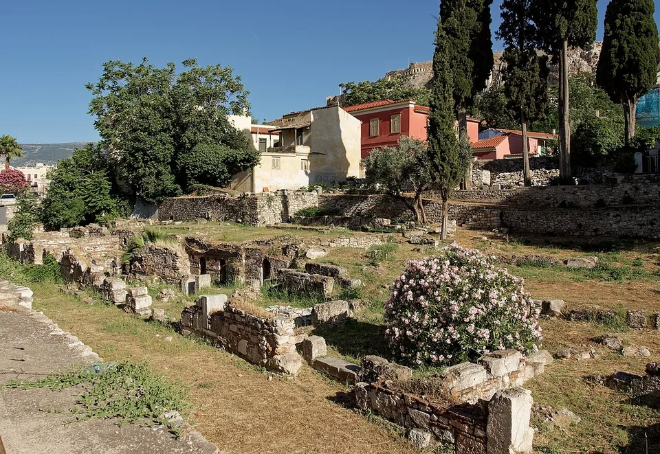 Low ruined walls and the bases of small rooms among grass and a flowering oleander, with houses of the Plaka and the Acropolis rock behind.