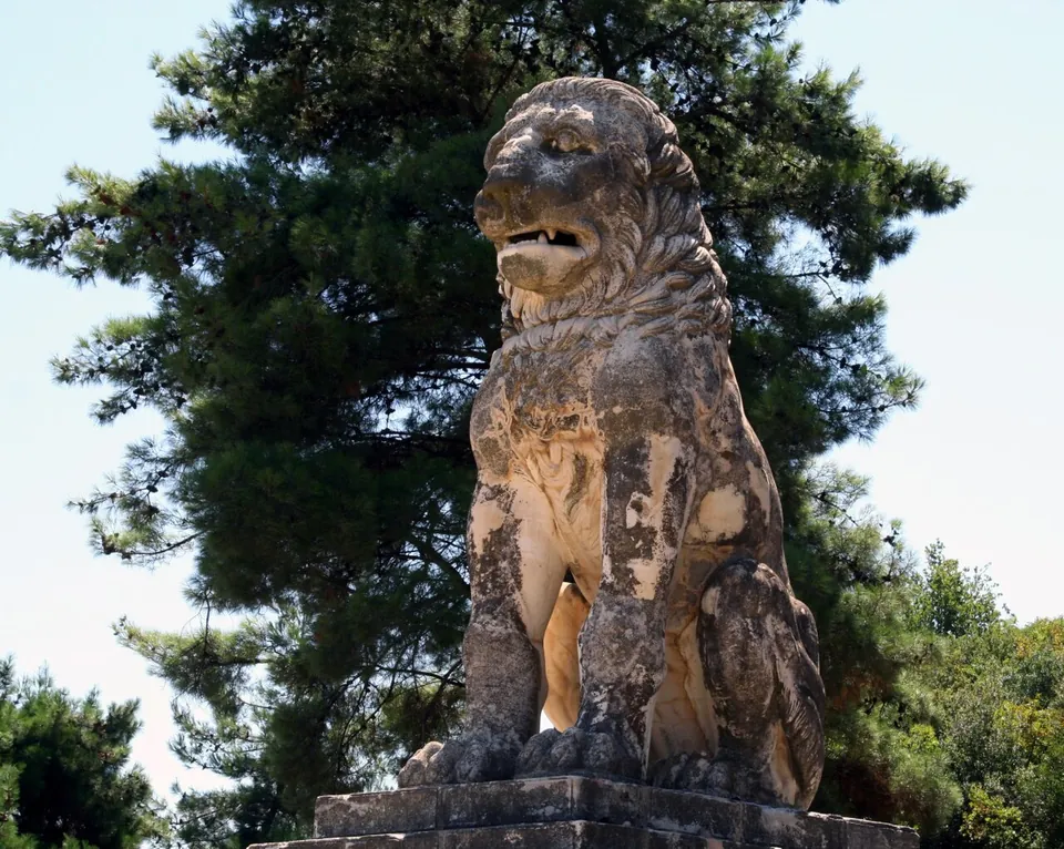 A colossal stone lion sitting upright on a plinth, its mouth open, in front of a tall pine against a pale sky.