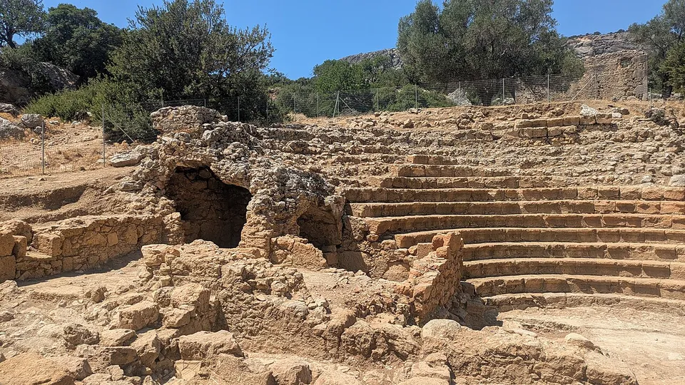 Curved rows of stone seats rising in a semicircle beside the broken vaults of a stone substructure, with olive trees and a wire fence behind.