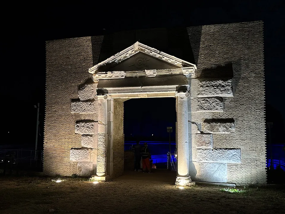 A portal with two columns and a pediment lit by floodlights at night, in a high wall of small stones with larger blocks set into it, the opening glowing blue from lighting beyond.