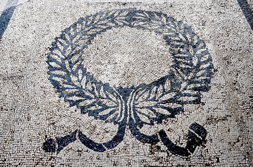 Close-up of a black-and-white mosaic floor of small stone cubes, showing a laurel wreath tied with a ribbon whose ends trail below it.