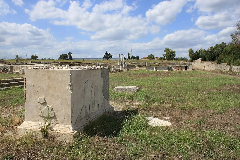 A large rectangular stone pedestal on a moulded plinth, with a few fragments of its marble facing set back onto its sides, stands in rough grass; behind it low walls, a few standing columns and a long boundary wall stretch across a flat, open site under a cloudy sky.