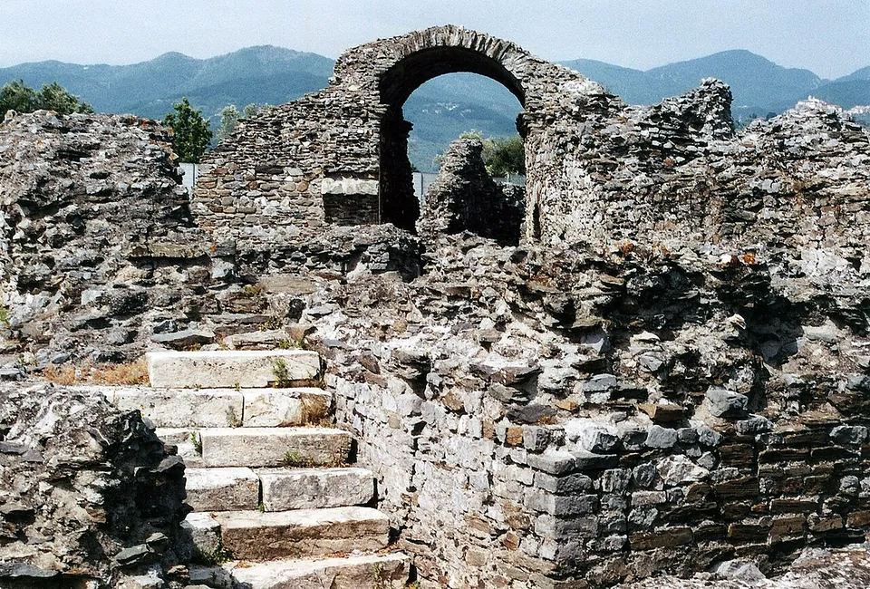 A round-headed arch of rough stone standing above broken walls, with a short flight of pale stone steps leading up in the foreground and wooded hills behind.