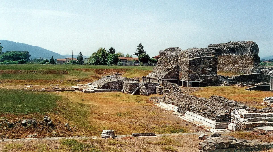 A broad grassy excavation with low walls and paving across the foreground and, to the right, a tall block of ruined rubble masonry, with trees, houses and a line of hills behind.
